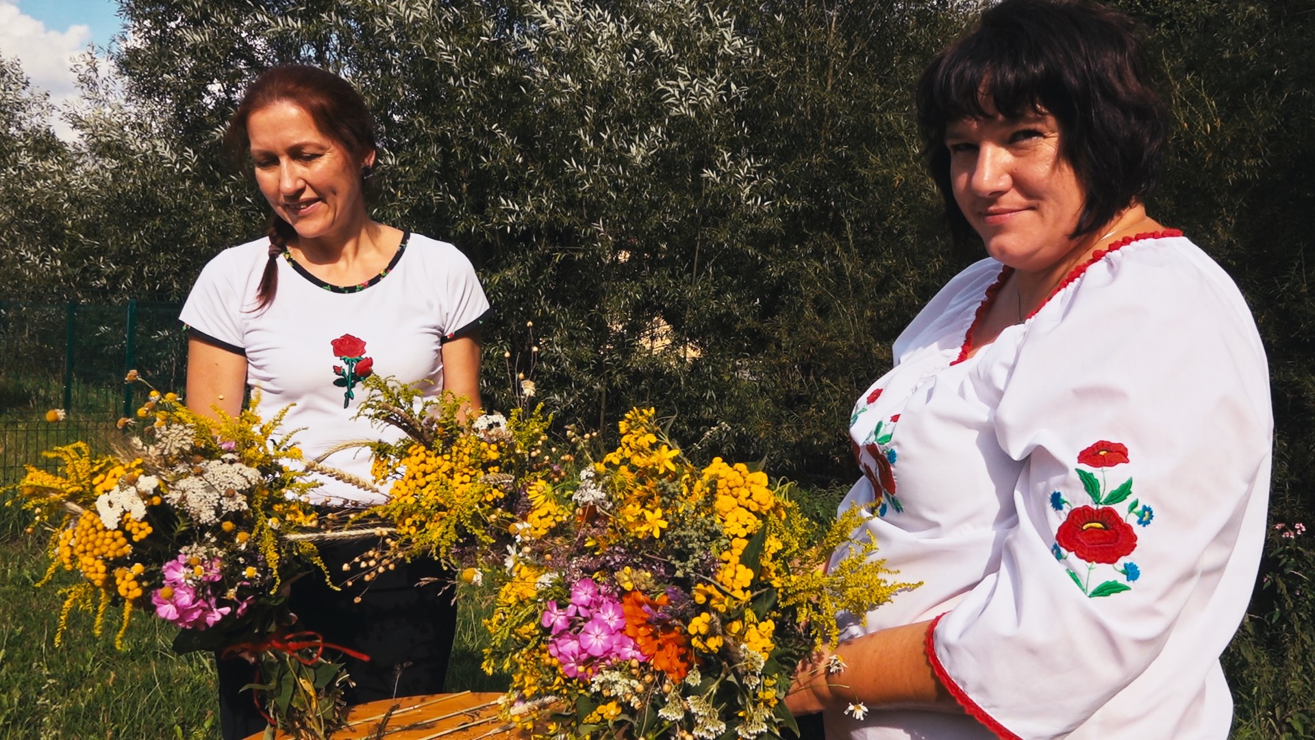dwie kobiety w białych bluzkach prezentują bukiety z żółtych, fioletowych i pomarańczowych kwiatów